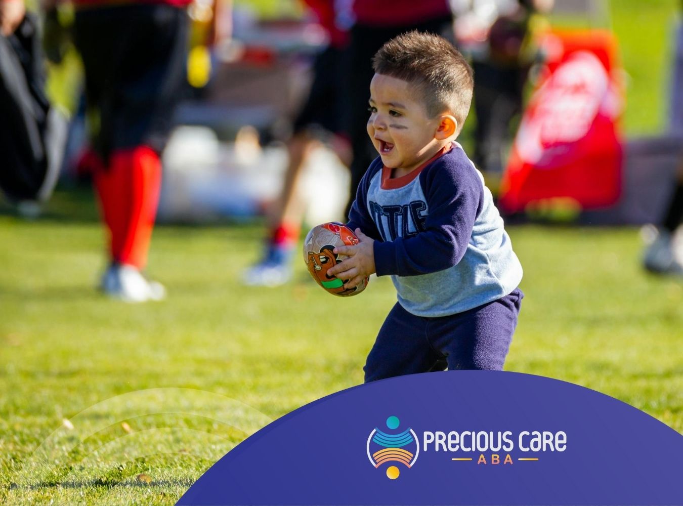 An autistic child excitedly holds a small football on a field, with players at the back in Colorado.