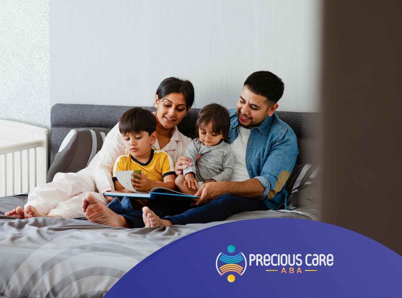A family of four sits on a bed reading together. The parents smile warmly, engaging their two young children with a book, creating a cozy, loving atmosphere.