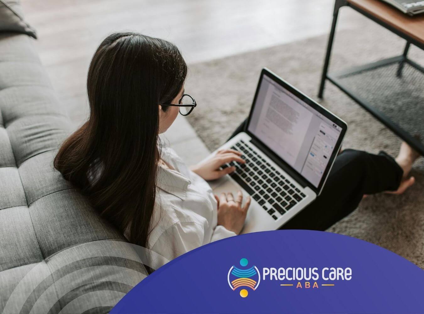 ABA therapy woman sitting on floor using a laptop beside a sofa, writing a report for Precious ABA.