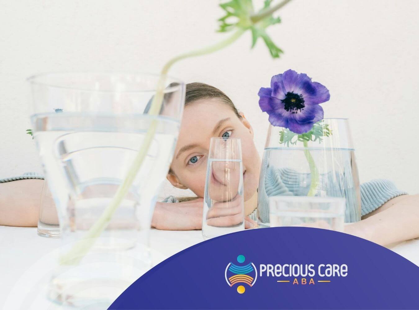 An Autistic woman looking through glasses of water with a purple flower on the table after ABA Therapy