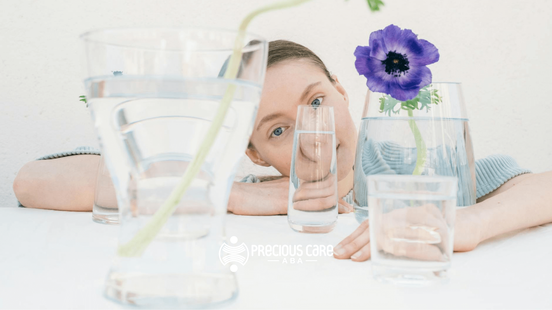 An Autistic woman looking through glasses of water with a purple flower on the table after ABA Therapy